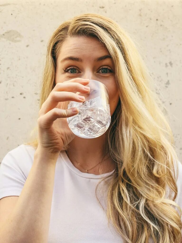 A woman with blonde hair and in a white t-shirt drinking a glass of water